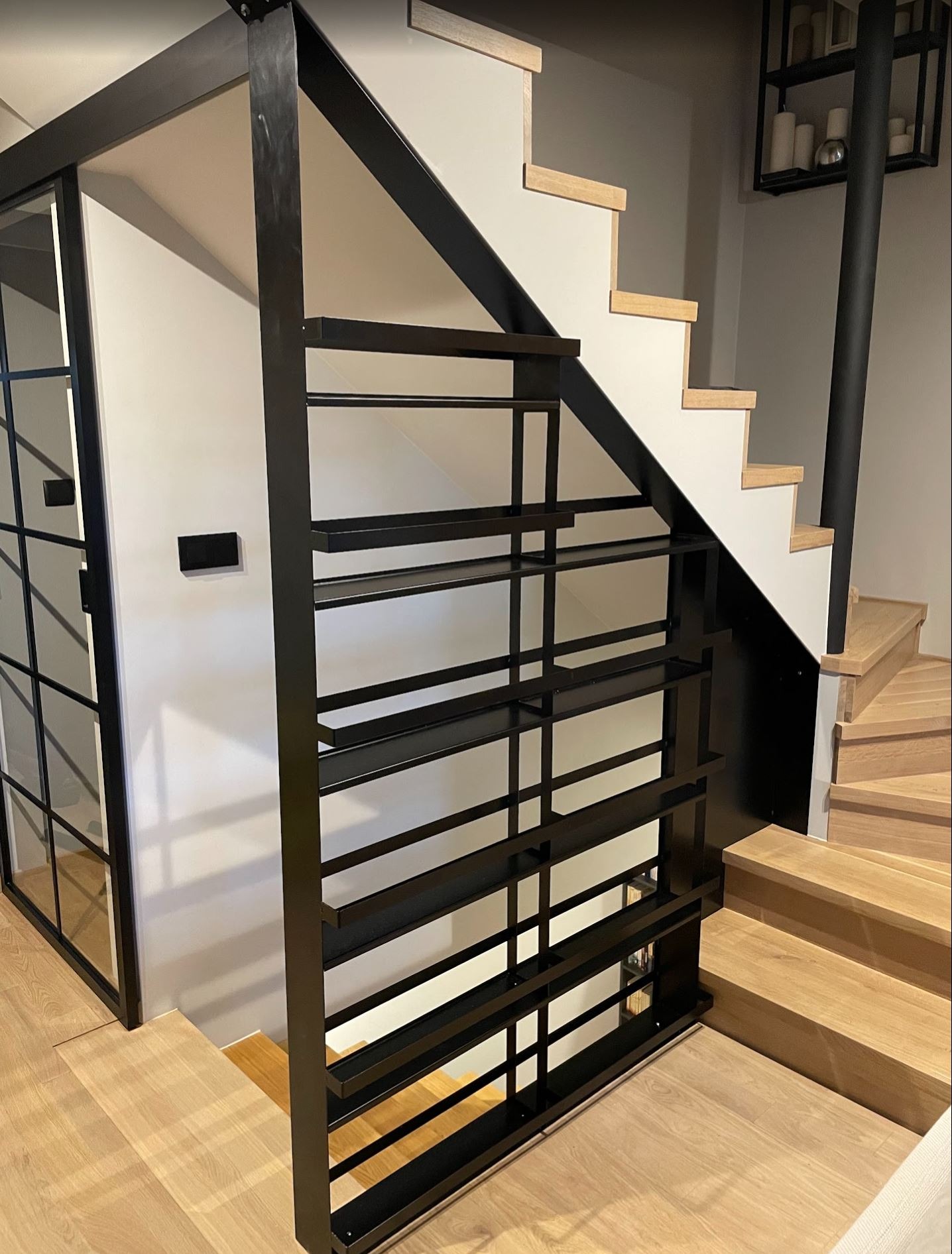 Ażurowy regał ze stali czarnej, domknięcie klatki schodowej z przesuwnymi drzwiami industrialnymi. Widoczne drewniane schody i czarna półka na ścianie.