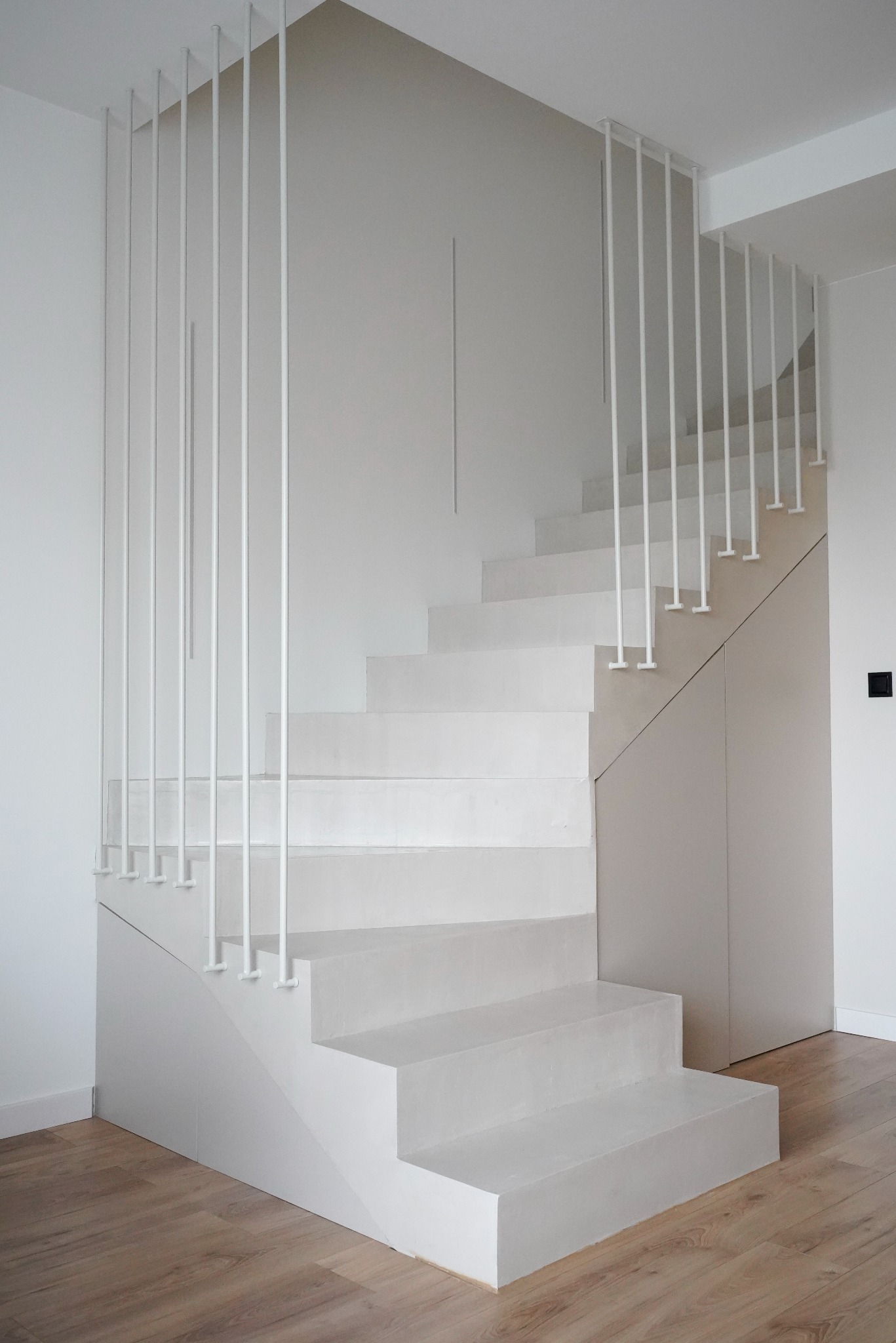 Minimalistyczne, białe schody z metalową balustradą z pionowych prętów w nowoczesnym domu. Widoczne drewniane panele podłogowe i fragment zabudowy pod schodami.