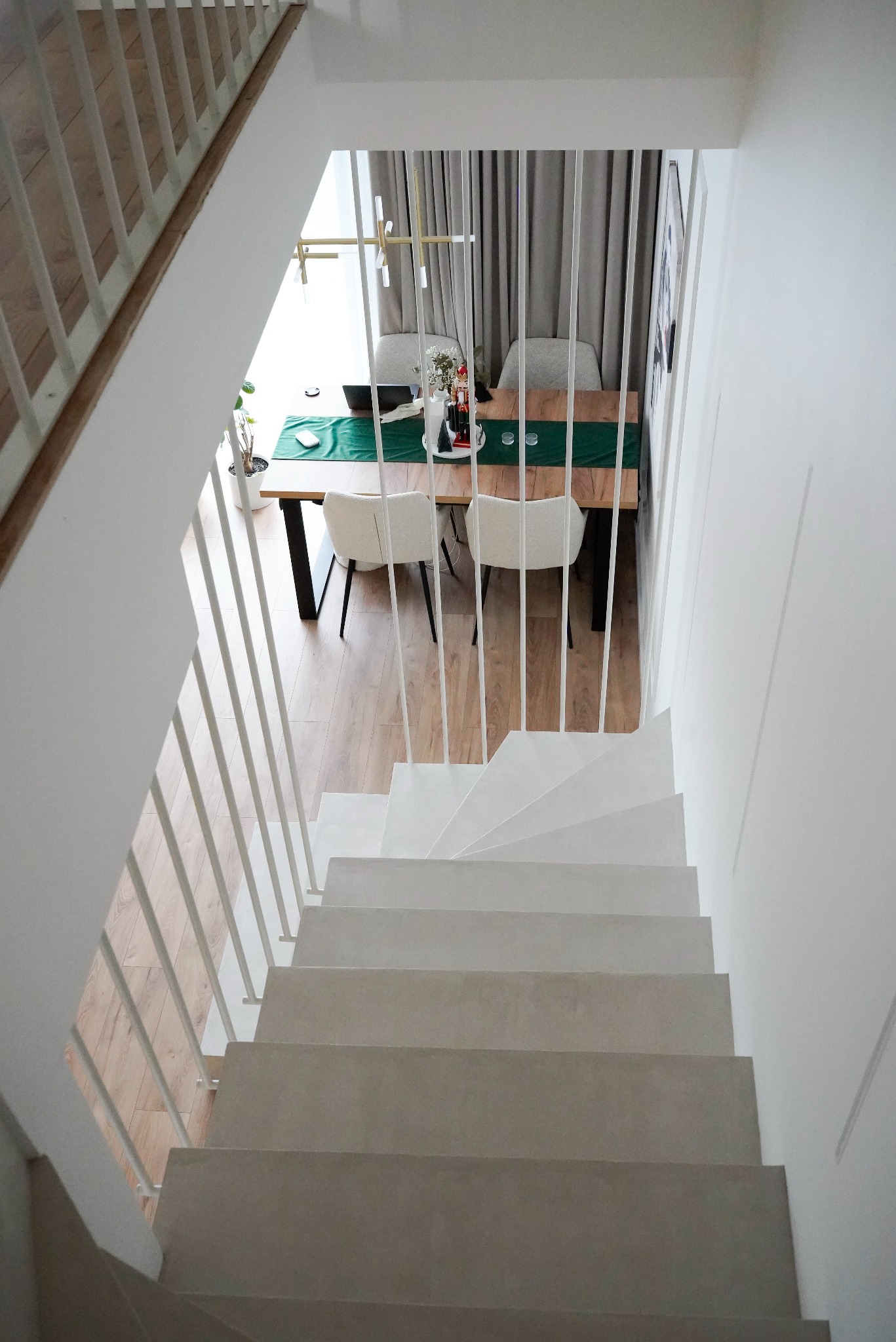Minimalistyczne schody z białą, metalową balustradą w nowoczesnym domu. Widok z góry na drewniany stół i krzesła w jadalni. Jasne, naturalne światło.
