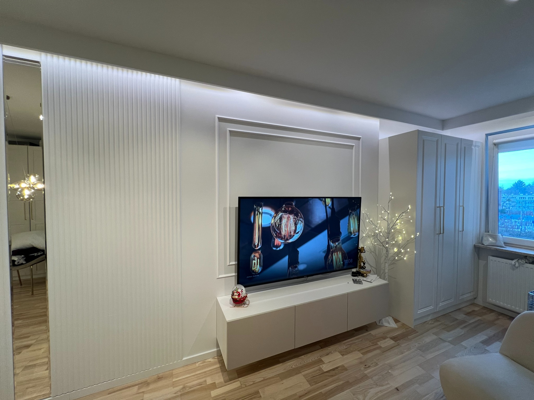 Nowoczesny salon z białą ścianą zdobioną sztukaterią i lamelami, podświetlony telewizor na szafce RTV, drewniana podłoga, okno z widokiem na miasto.