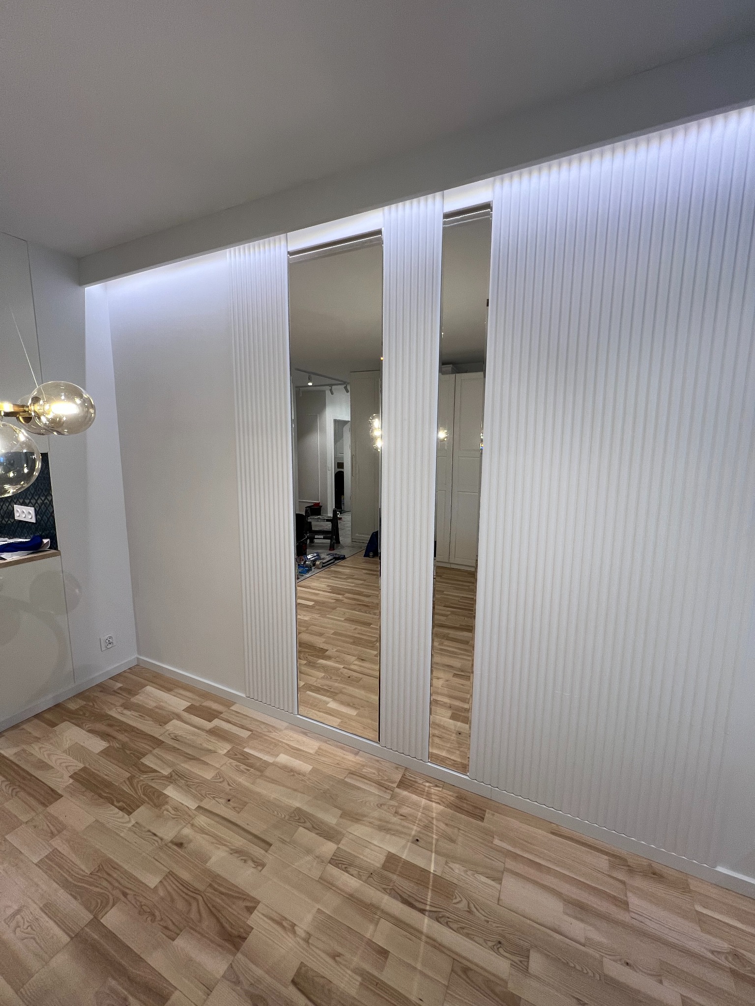 Nowoczesna szafa wnękowa z lustrzanymi drzwiami przesuwnymi i dekoracyjnymi panelami ściennymi. Parkiet na podłodze, oświetlenie LED na suficie.