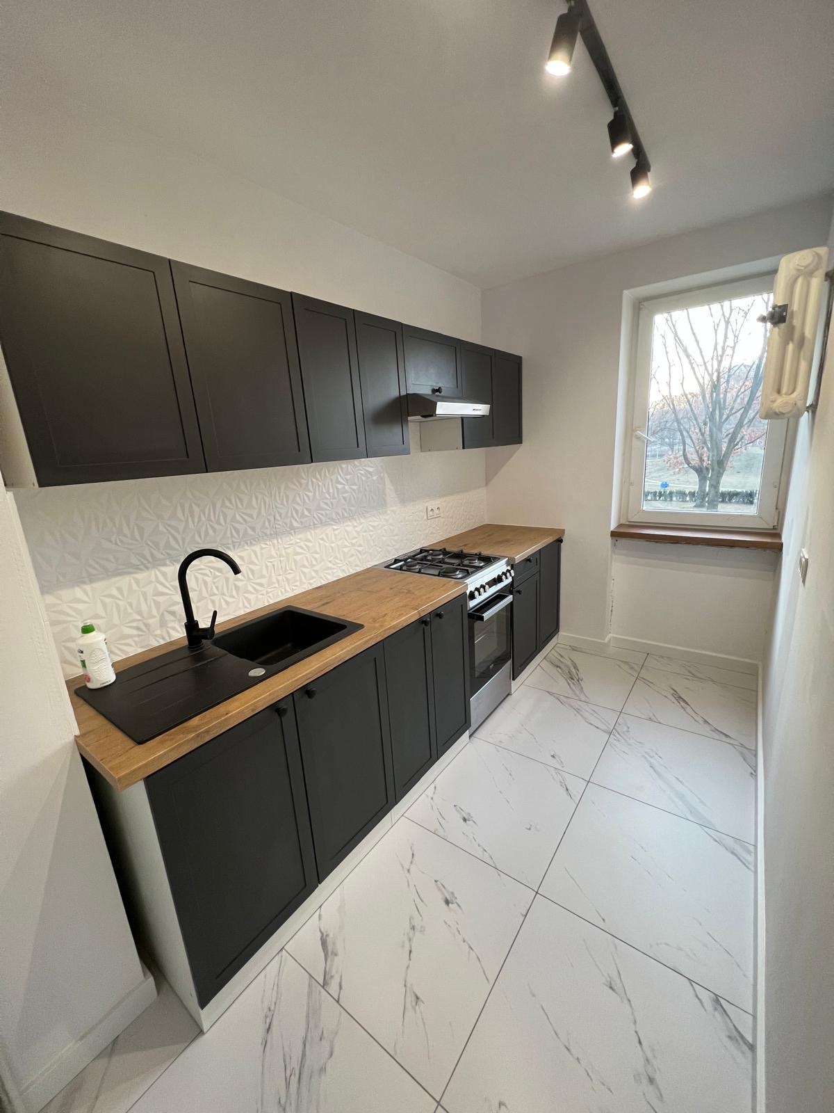 Nowoczesna kuchnia z czarnymi szafkami, drewnianym blatem i białymi płytkami z geometrycznym wzorem. Widok na okno z drzewem. Oświetlenie punktowe.