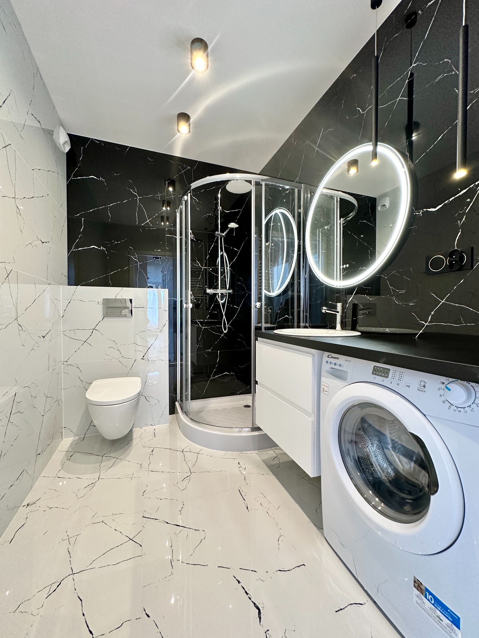 Nowoczesna łazienka w czarno-białej kolorystyce z kabiną prysznicową, toaletą, umywalką z podświetlanym lustrem i pralką. Marmurowe płytki na ścianach i podłodze.