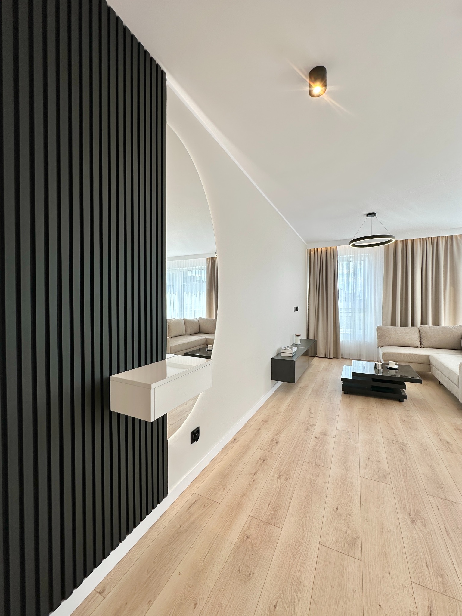 Nowoczesne wykończenie wnętrza: czarne lamele na ścianie, lustro z półką, jasna podłoga i salon w tle. Minimalistyczny design z dbałością o detale.