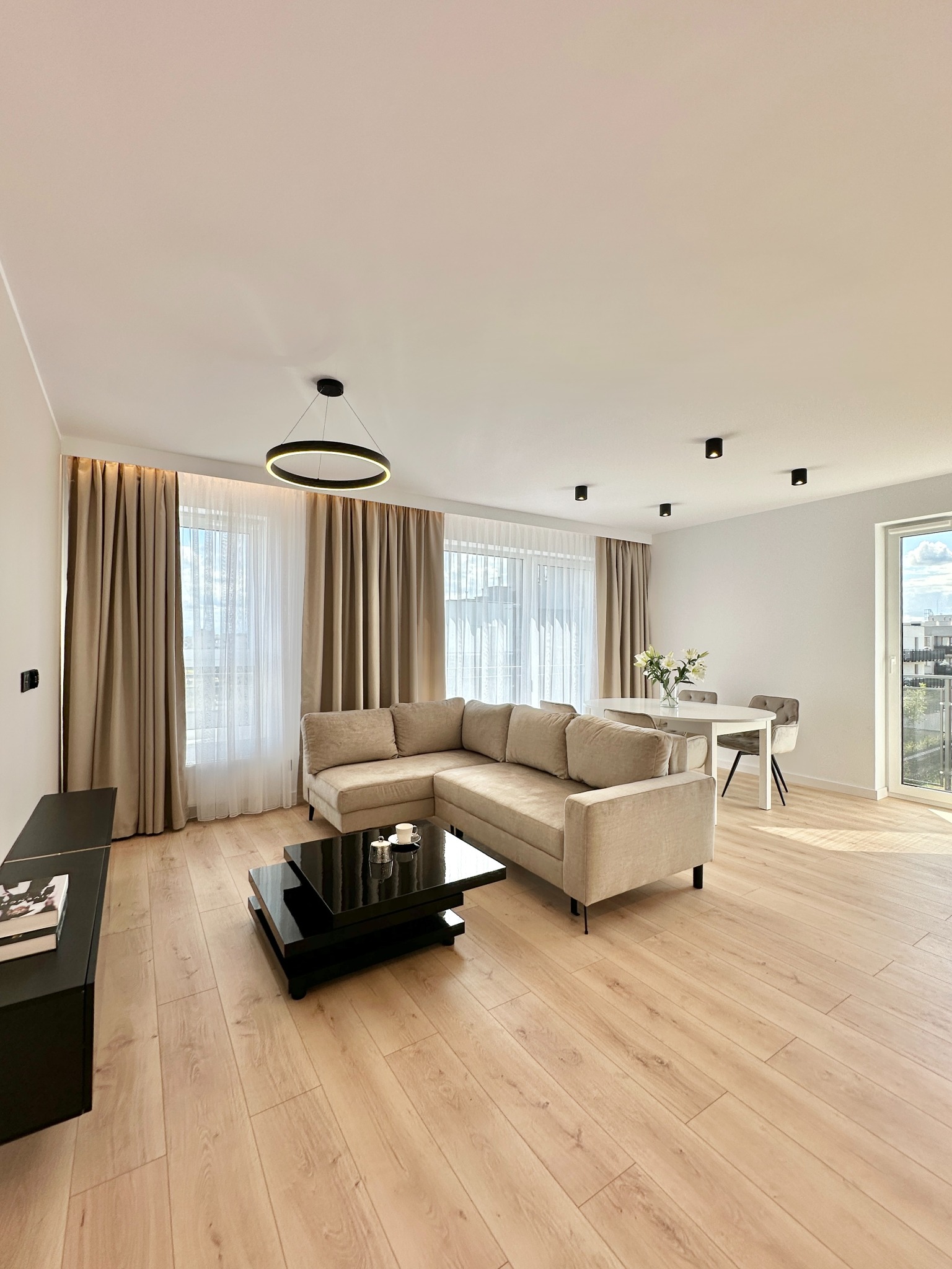 Jasne wnętrze salonu z beżową sofą narożną, czarnym stolikiem kawowym i parkietem. Okno z zasłonami w odcieniach beżu. Nowoczesne oświetlenie sufitowe.