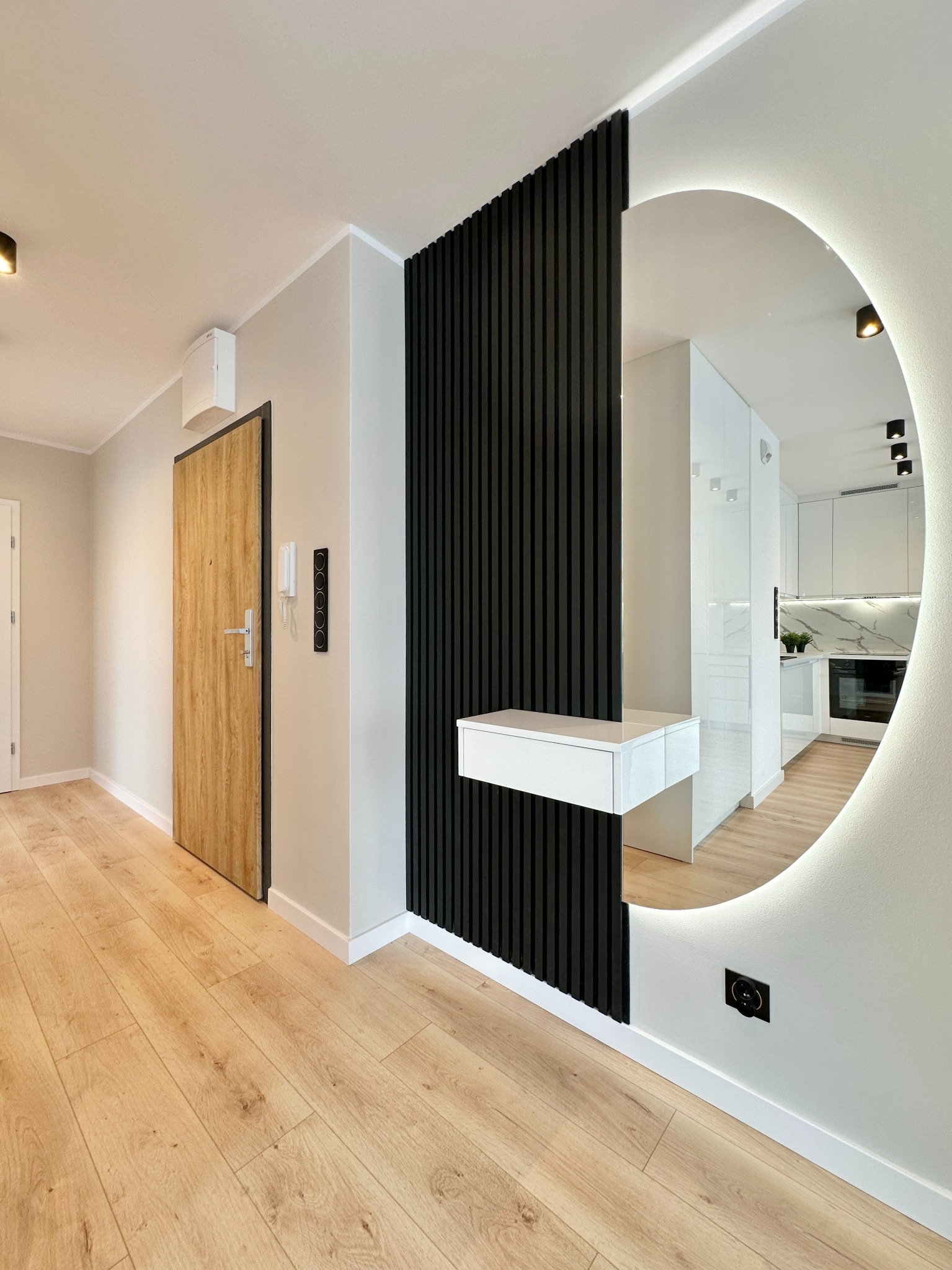 Nowoczesny przedpokój z drewnianą podłogą, drzwiami i czarną ścianką z lamelami. Okrągłe lustro z podświetleniem odbija wnętrze kuchni.