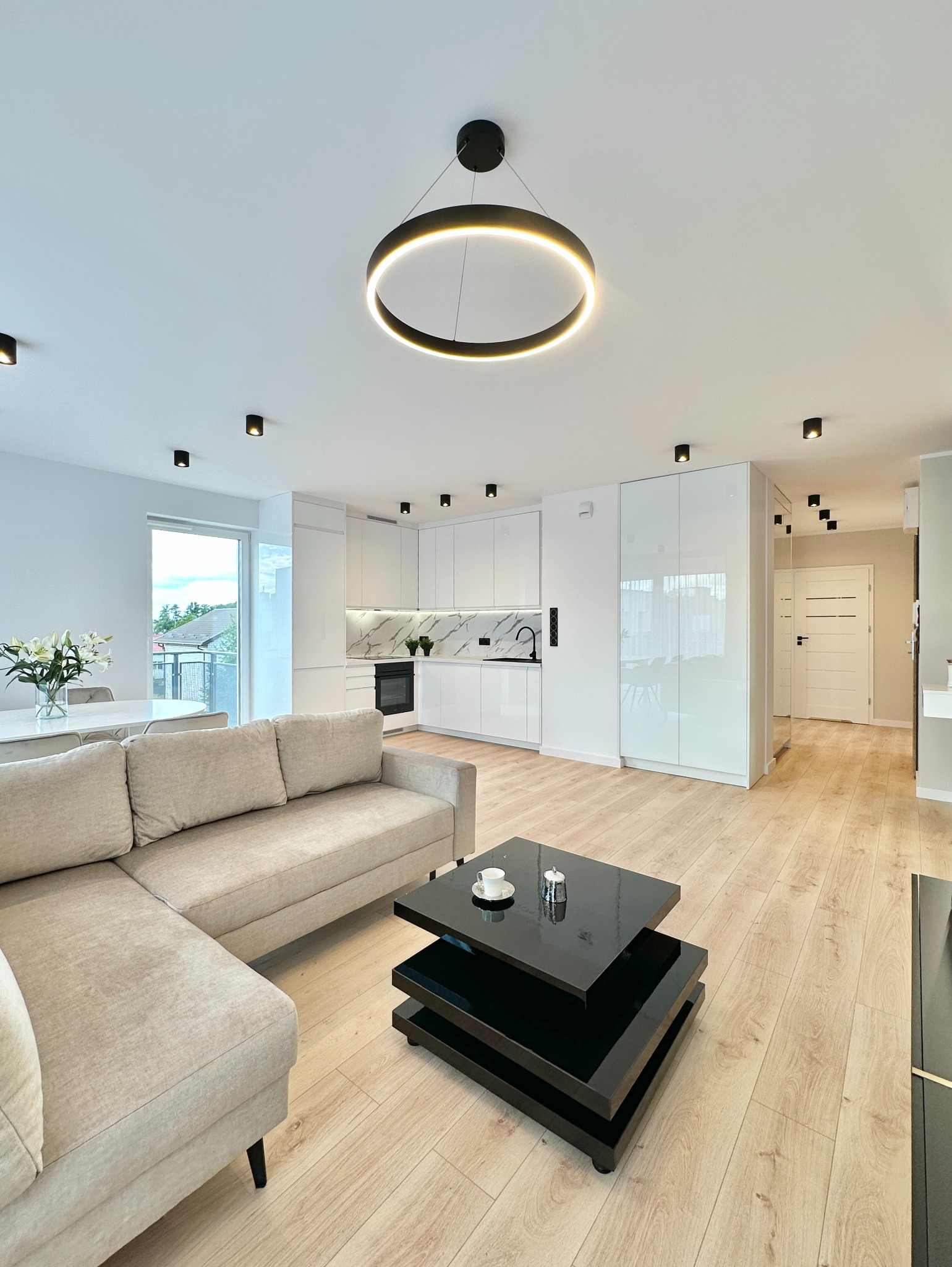 Jasne, nowoczesne wnętrze: beżowa sofa narożna, czarny stolik kawowy, biała kuchnia z marmurowym backsplashiem i okrągła lampa sufitowa. Minimalistyczny design.