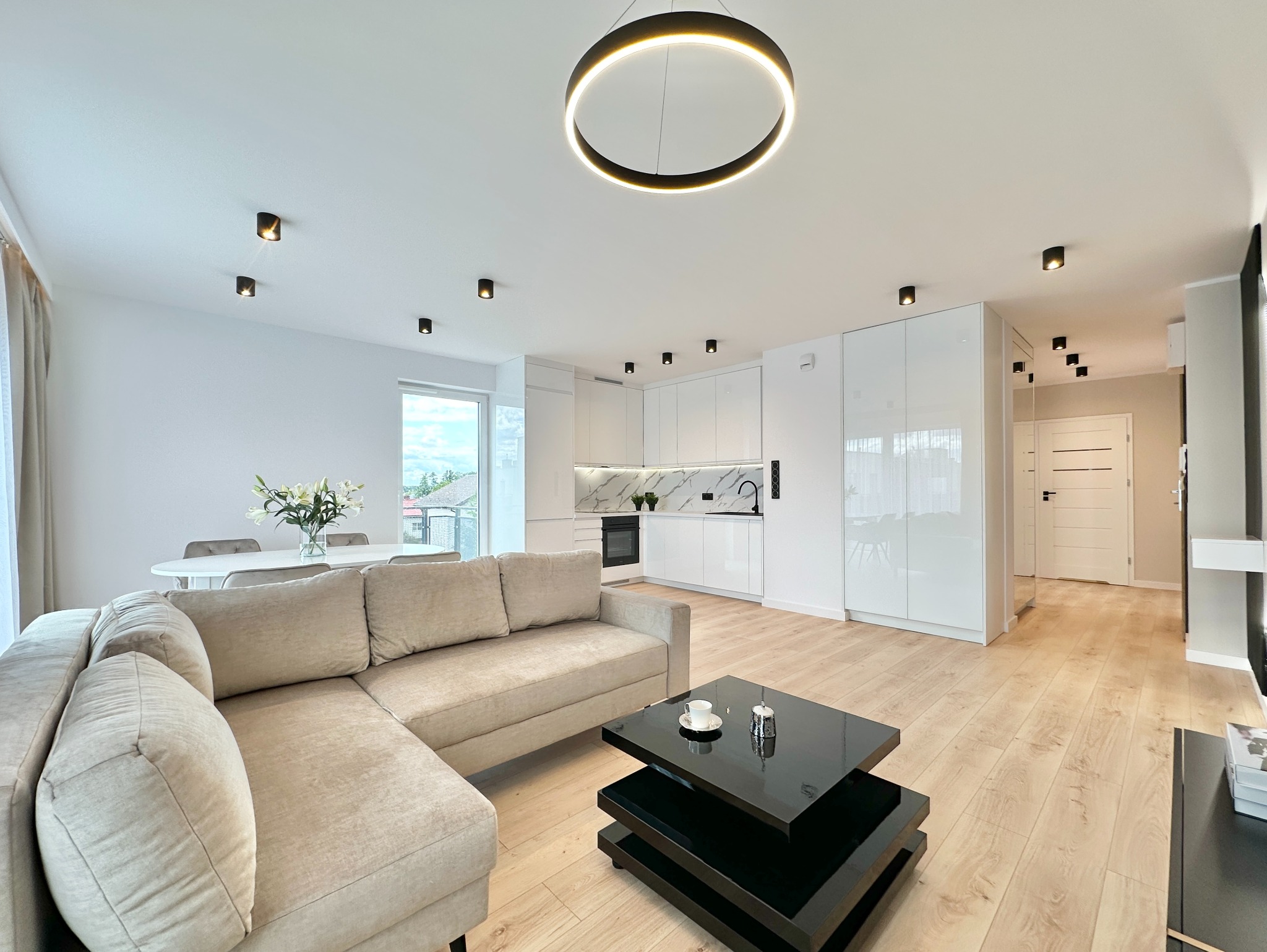 Jasne, przestronne wnętrze po remoncie: beżowa sofa narożna, czarny stolik kawowy, biała kuchnia z marmurowym blatem, drewniana podłoga i okrągła lampa sufitowa.