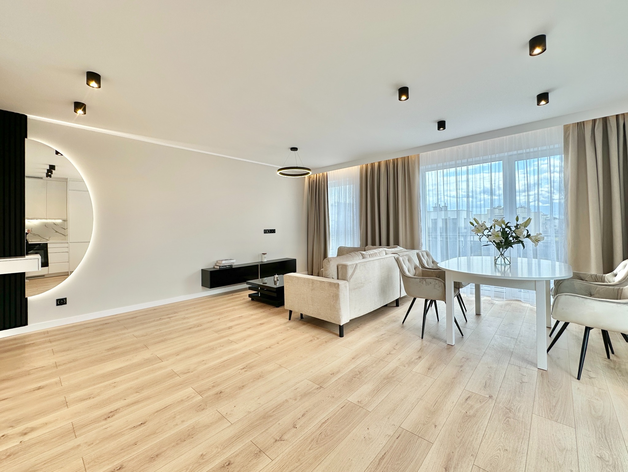 Jasne, przestronne wnętrze salonu z drewnianą podłogą, okrągłym lustrem z podświetleniem i nowoczesnym oświetleniem sufitowym. Minimalistyczny design.