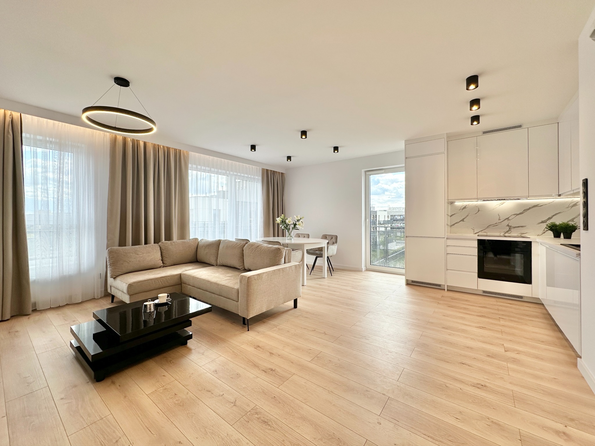 Jasne, przestronne wnętrze: parkiet, beżowa sofa narożna, czarny stolik kawowy, biała kuchnia z marmurowym blatem, okrągła lampa. Nowoczesny design.
