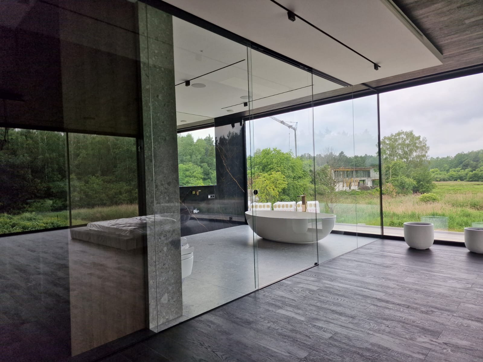 Nowoczesna łazienka z panoramicznym oknem i szklaną ścianą działową, widok na zieleń. Minimalistyczny design, wolnostojąca wanna, ciemna podłoga i marmurowe akcenty.