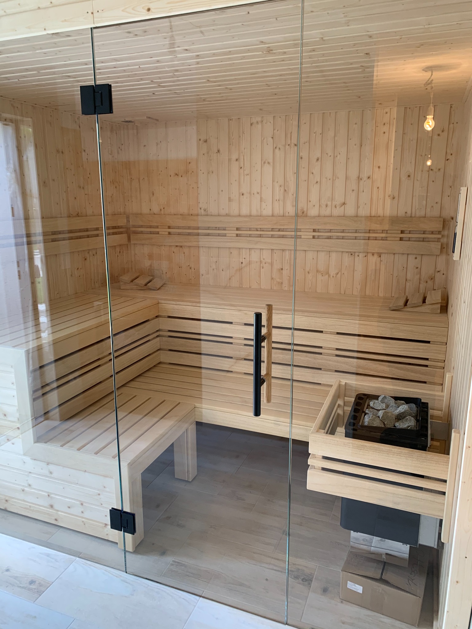 Jasne drewno i szkło tworzą nowoczesną saunę. Widoczne ławy, piec z kamieniami i szklane drzwi z czarnymi zawiasami. Minimalistyczny design i relaksująca atmosfera.