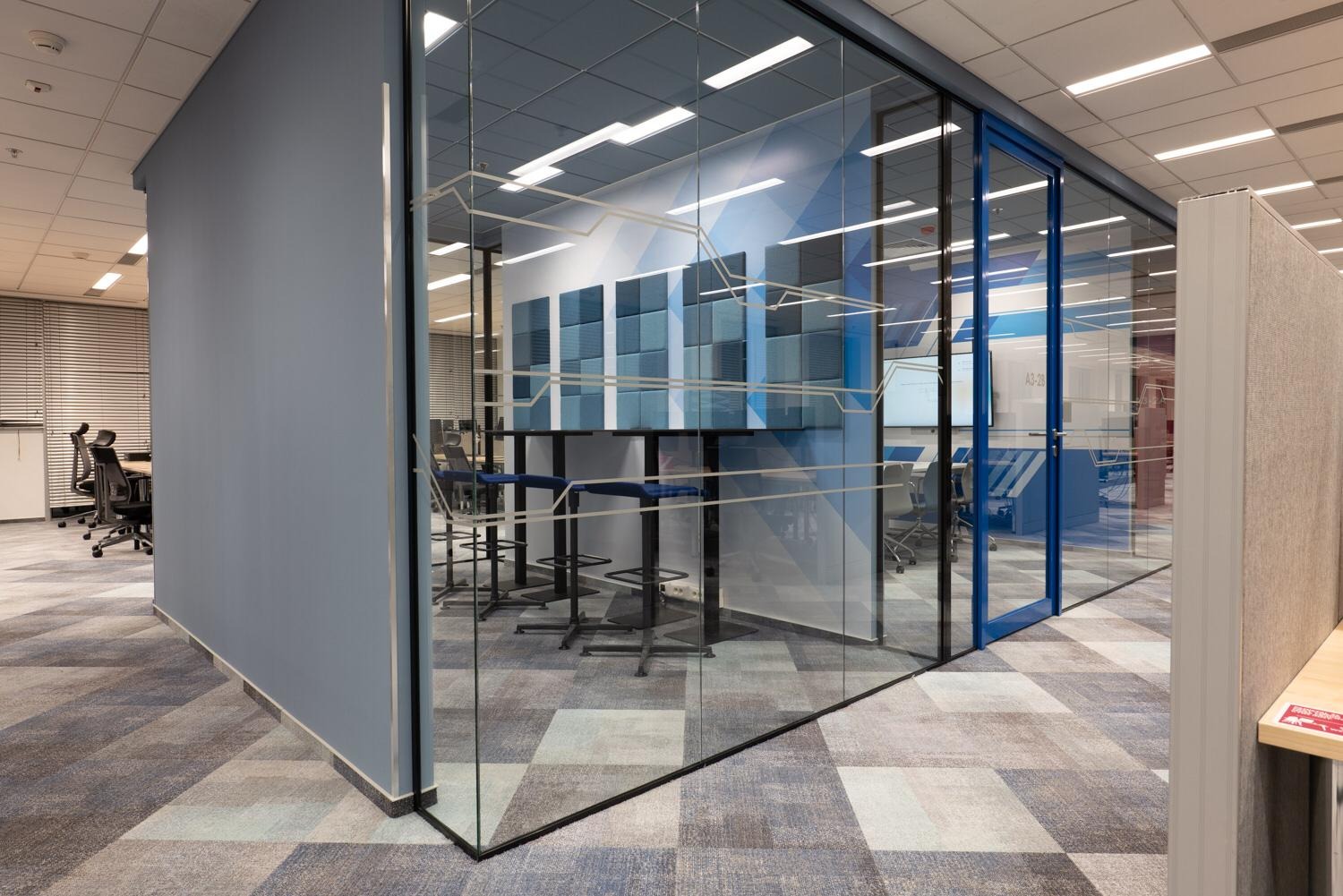 Szklana ścianka działowa z grafiką w biurze. Widoczne krzesła barowe, stół i elementy wystroju w odcieniach szarości i błękitu. Nowoczesne wnętrze biurowe.