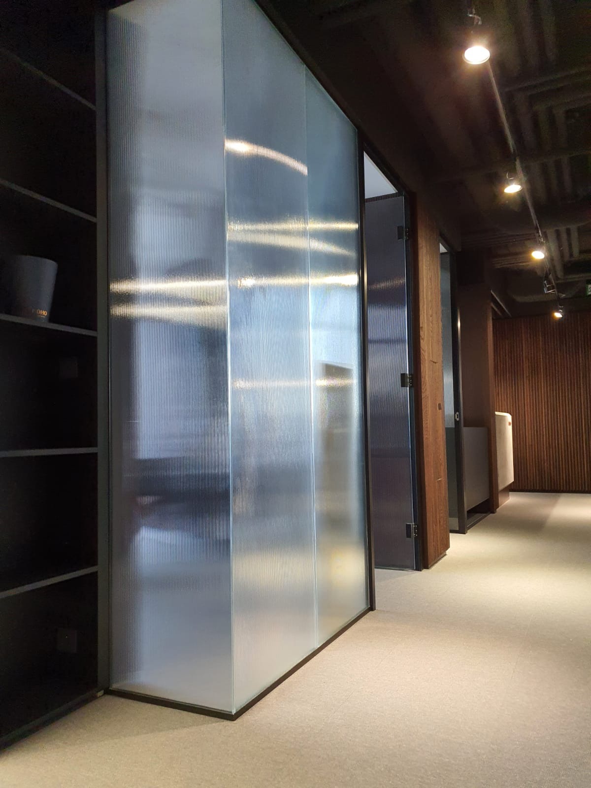 Szklana ścianka działowa z ryflowanego szkła w czarnej ramie, oddzielająca przestrzeń biurową. Widoczne oświetlenie sufitowe i fragmenty wyposażenia.