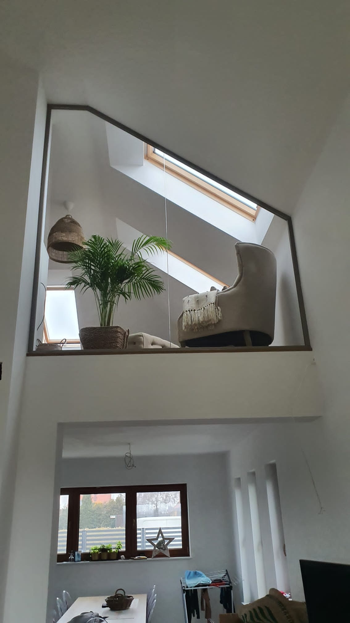 Dwupoziomowe wnętrze z antresolą zabezpieczoną ramą, na której stoi fotel i roślina doniczkowa. W tle okno dachowe i widok na niższy poziom z oknem.