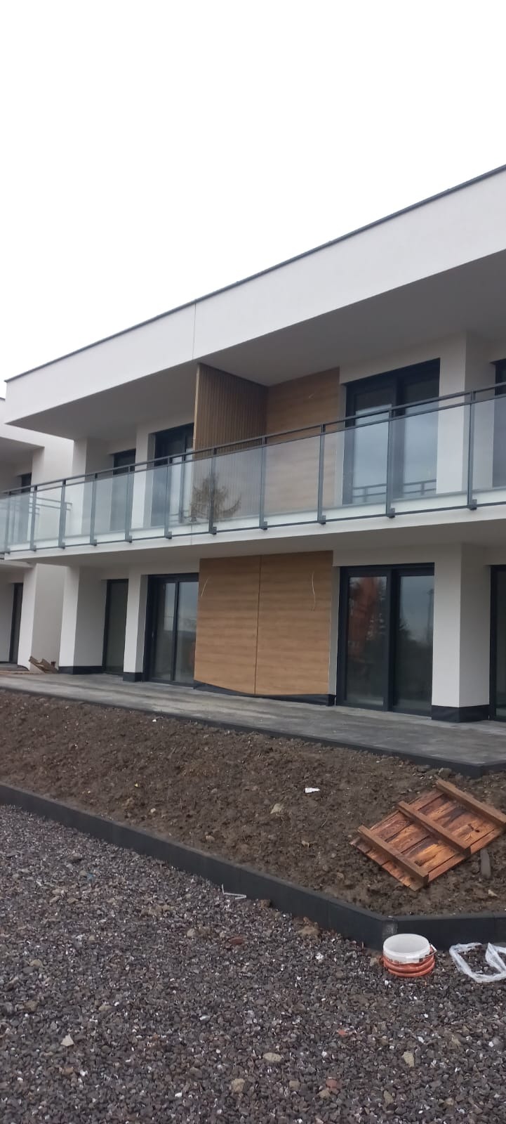 Nowoczesny budynek z balkonami zabezpieczonymi szkłem, widok z zewnątrz. Na pierwszym planie ziemia i gruz.