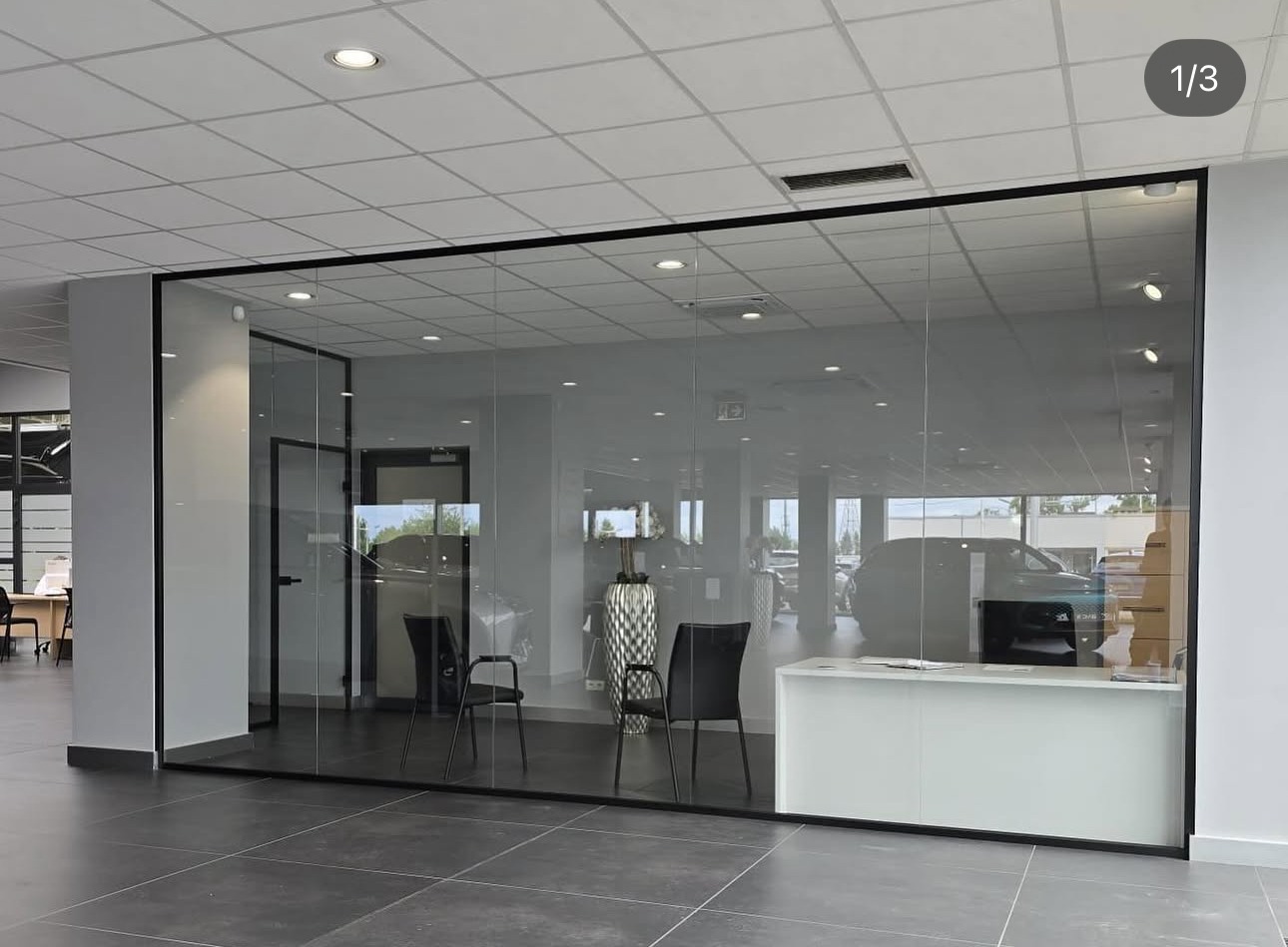 Szklana ściana działowa z czarną ramą oddzielająca przestrzeń biurową, widoczne krzesła i biurko w tle, szara podłoga z płytek.
