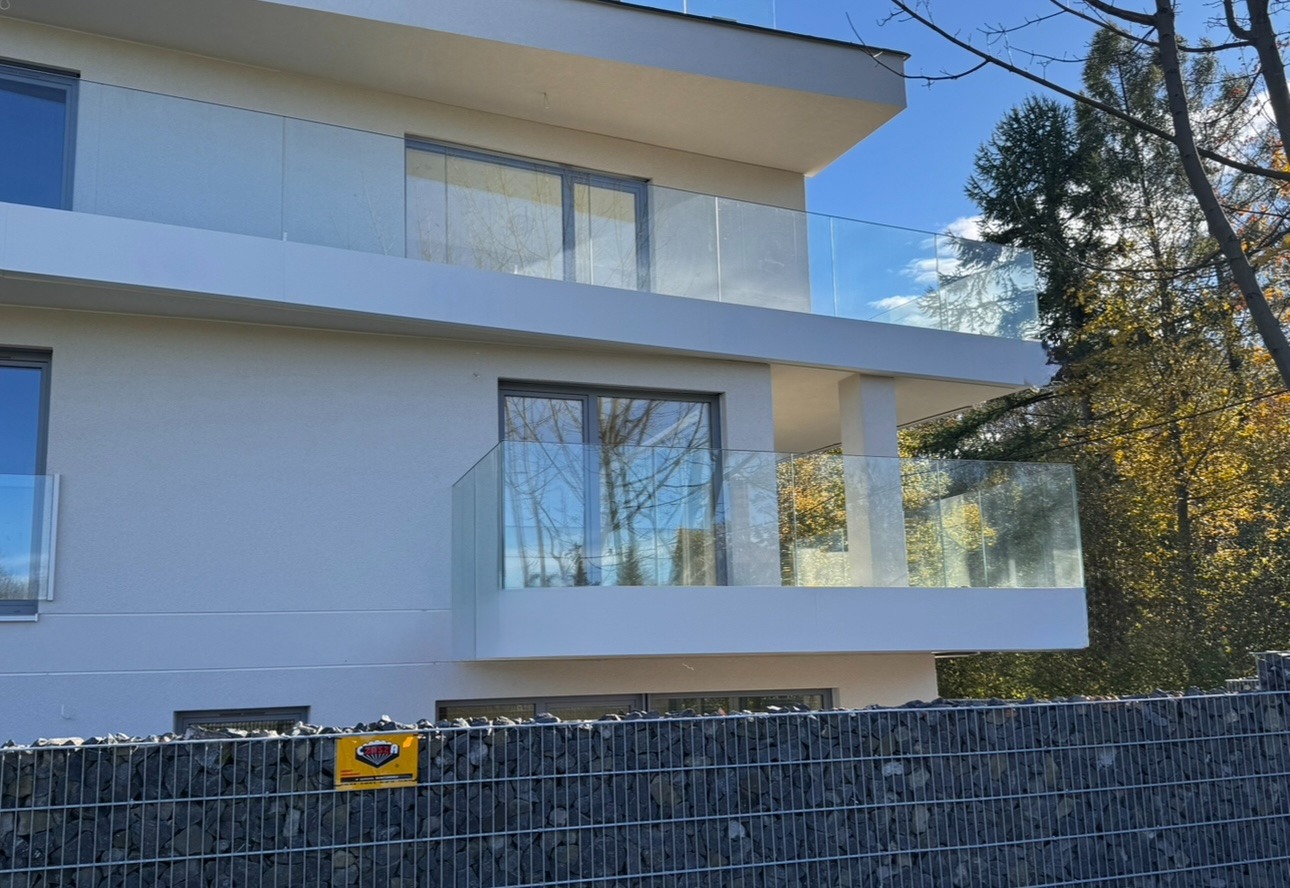 Nowoczesny budynek z balkonami zabezpieczonymi szkłem, widoczne refleksy drzew w szybach. Gabionowe ogrodzenie z żółtą tabliczką u dołu kadru.