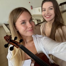 Julia Olczyk Skrzypce - Dwie uśmiechnięte kobiety, blondynka z przodu trzyma skrzypce, w tle jasne wnętrze z ławkami, możliwe sala koncertowa lub kościół. Selfie.