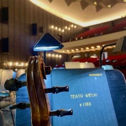 Julia Olczyk Skrzypce - Główka wiolonczeli na tle sceny Teatru Wielkiego, z widocznymi rzędami czerwonych foteli i oświetleniem. Nastrojowe oświetlenie sceniczne.