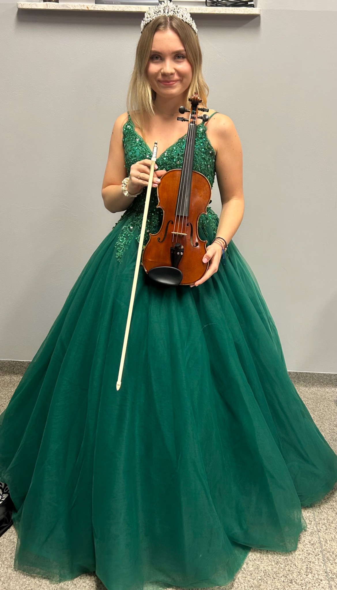Uśmiechnięta blondynka w zielonej sukni balowej i koronie trzyma skrzypce ze smyczkiem na tle szarej ściany. Strój sceniczny.