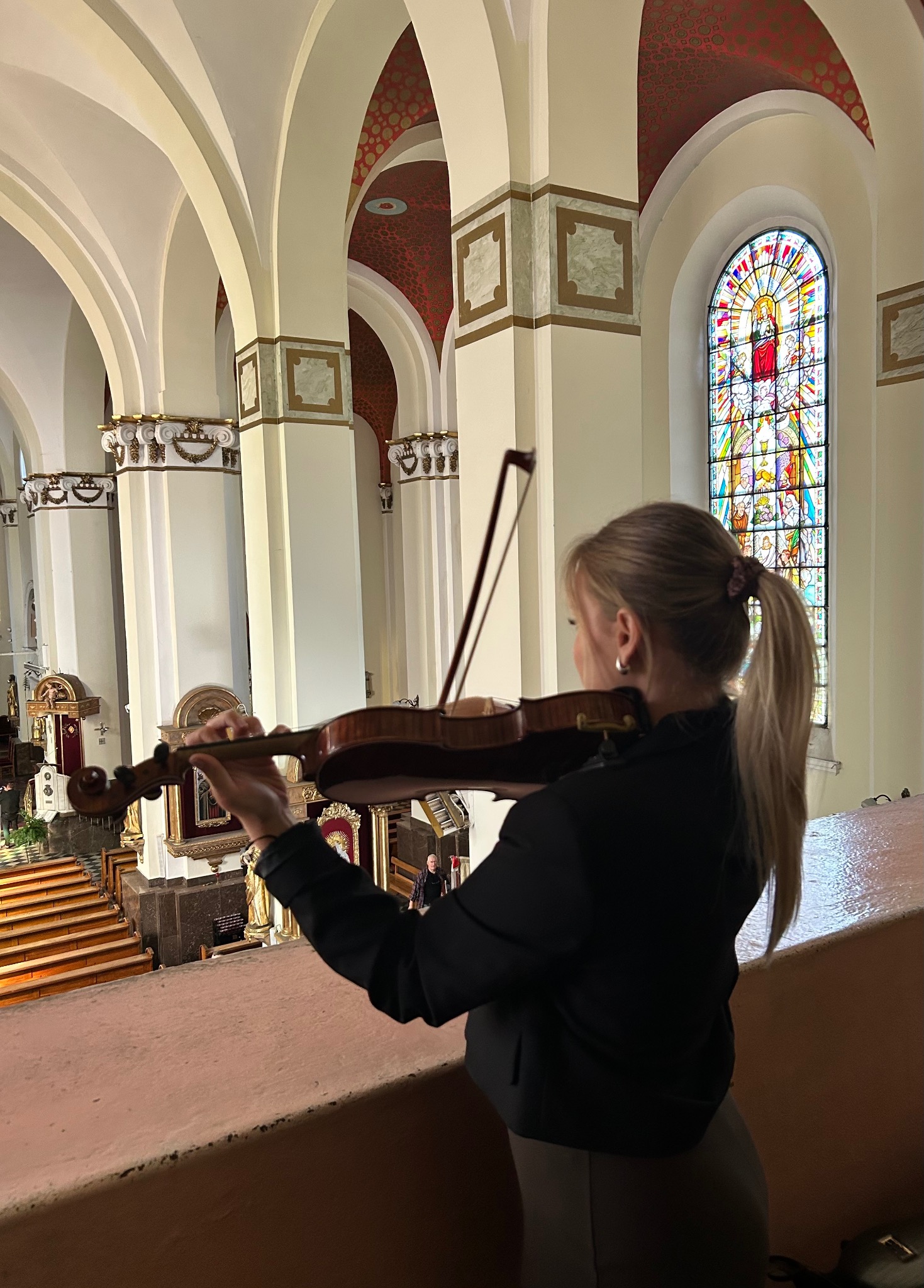 Skrzypaczka grająca w kościele z witrażem. Widok z góry na wnętrze świątyni, kolumny i łuki. Artystka w czarnej marynarce, z blond włosami spiętymi w kucyk.