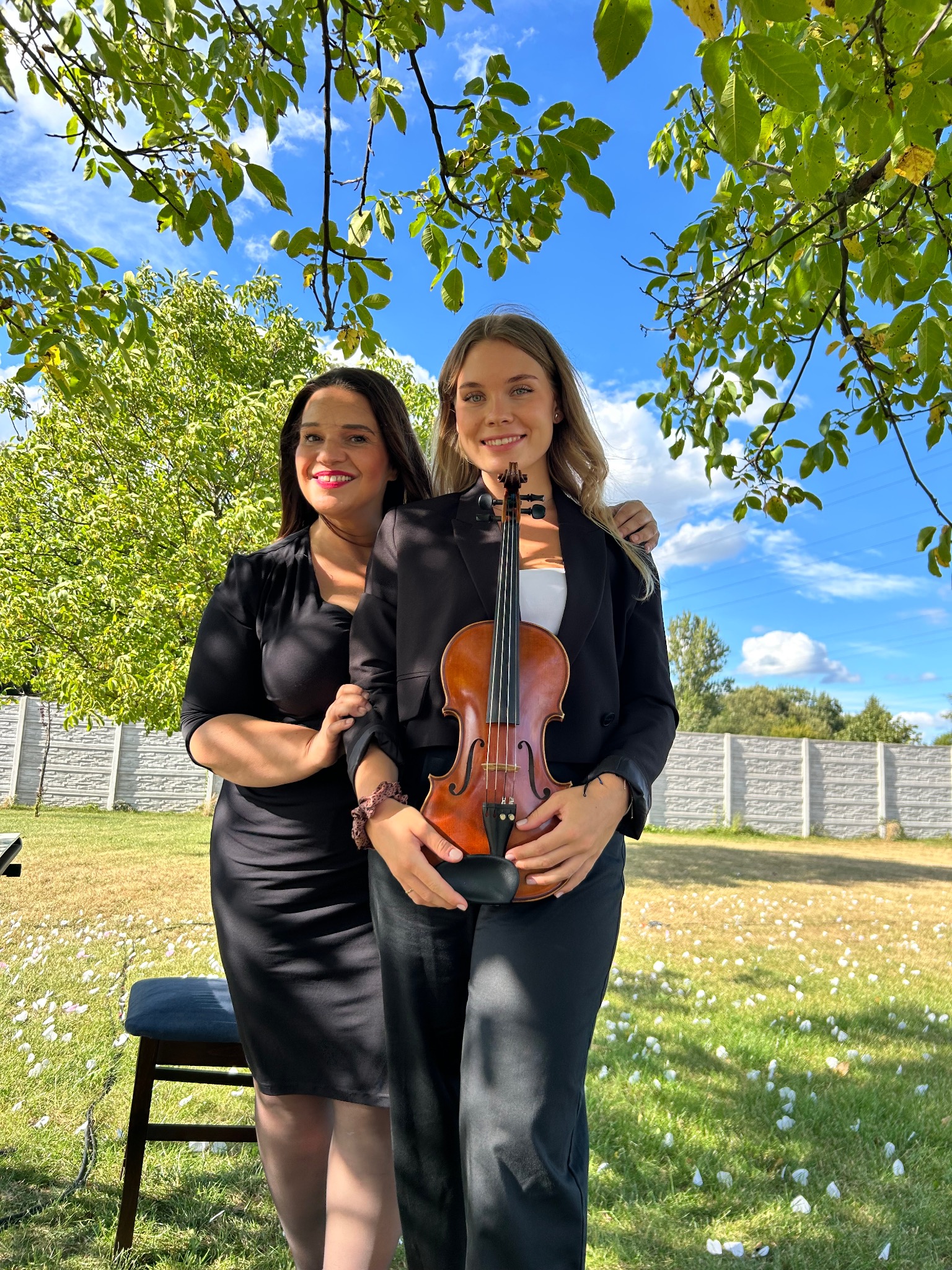 Dwie uśmiechnięte kobiety w plenerze, jedna trzyma skrzypce. W tle zieleń drzew i błękitne niebo z chmurami. Profesjonalny duet muzyczny na tle białego płotu.