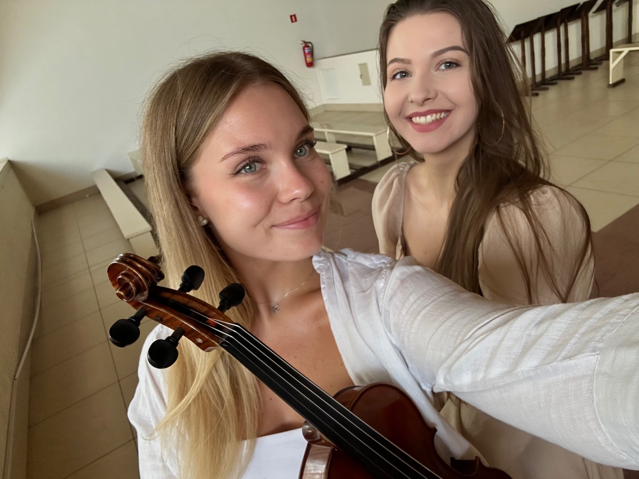 Dwie uśmiechnięte kobiety, blondynka z przodu trzyma skrzypce, w tle jasne wnętrze z ławkami, możliwe sala koncertowa lub kościół. Selfie.