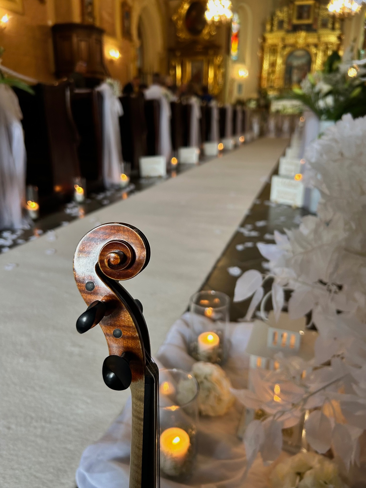 Skrzypce na tle kościoła udekorowanego na ślub. W tle ławki z białymi dekoracjami, świece i kwiaty. Ujęcie z bliska na główkę skrzypiec.