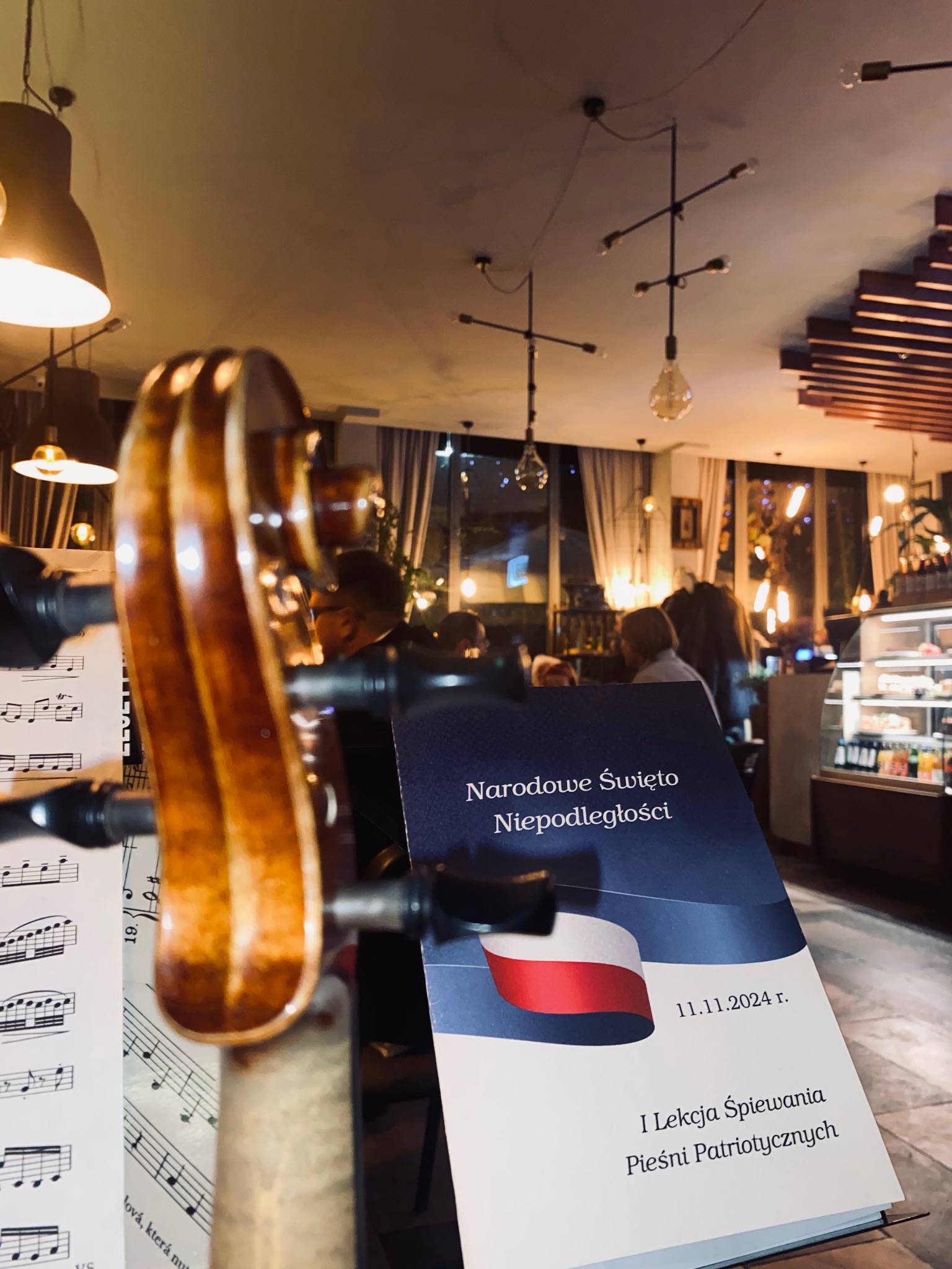 Skrzypce na tle sali z plakatem 'Narodowe Święto Niepodległości', nuty obok. Ciepłe światło lamp, w tle osoby w kawiarni. Ujęcie z bliska, perspektywa z dołu.