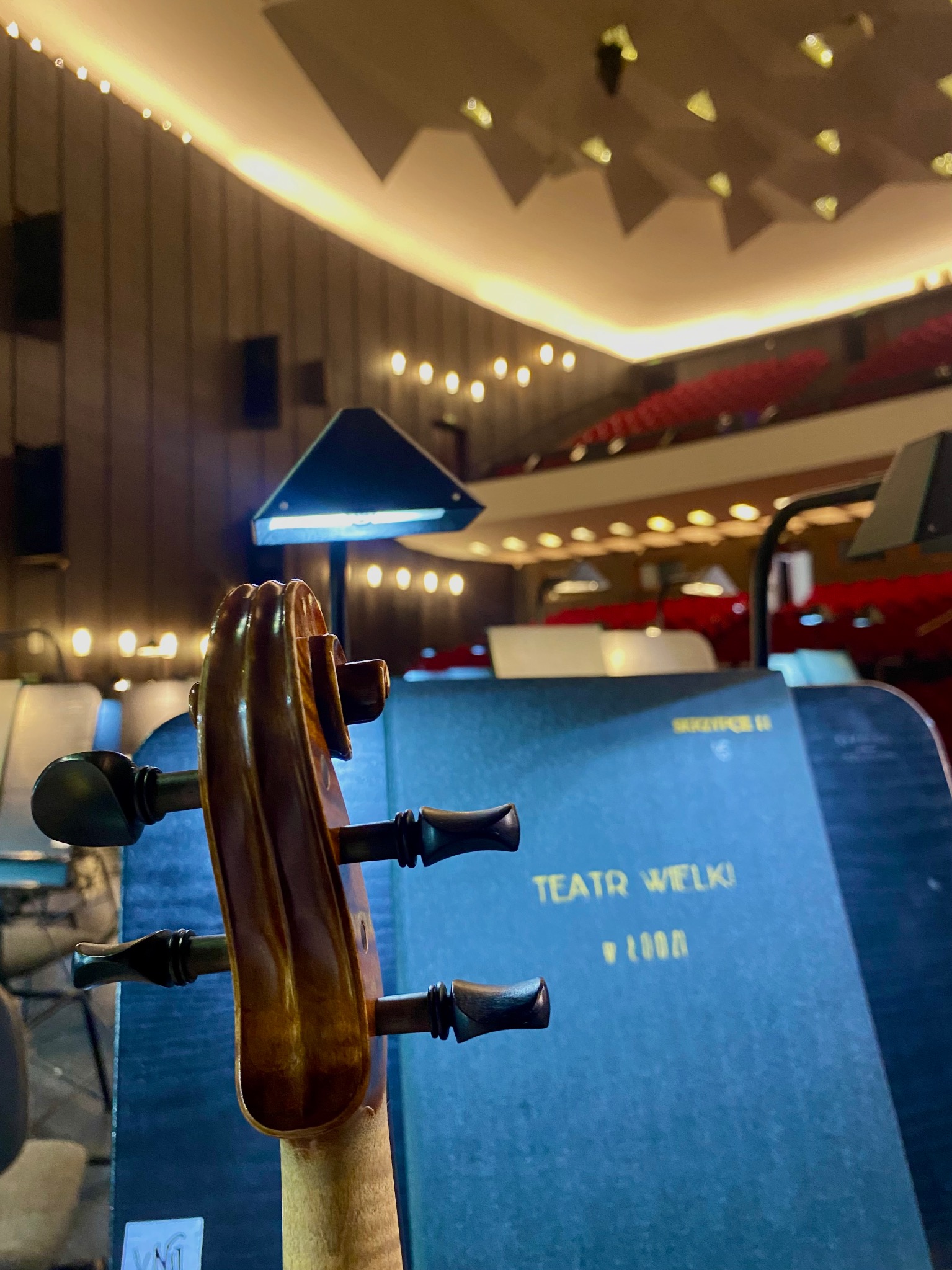 Główka wiolonczeli na tle sceny Teatru Wielkiego, z widocznymi rzędami czerwonych foteli i oświetleniem. Nastrojowe oświetlenie sceniczne.