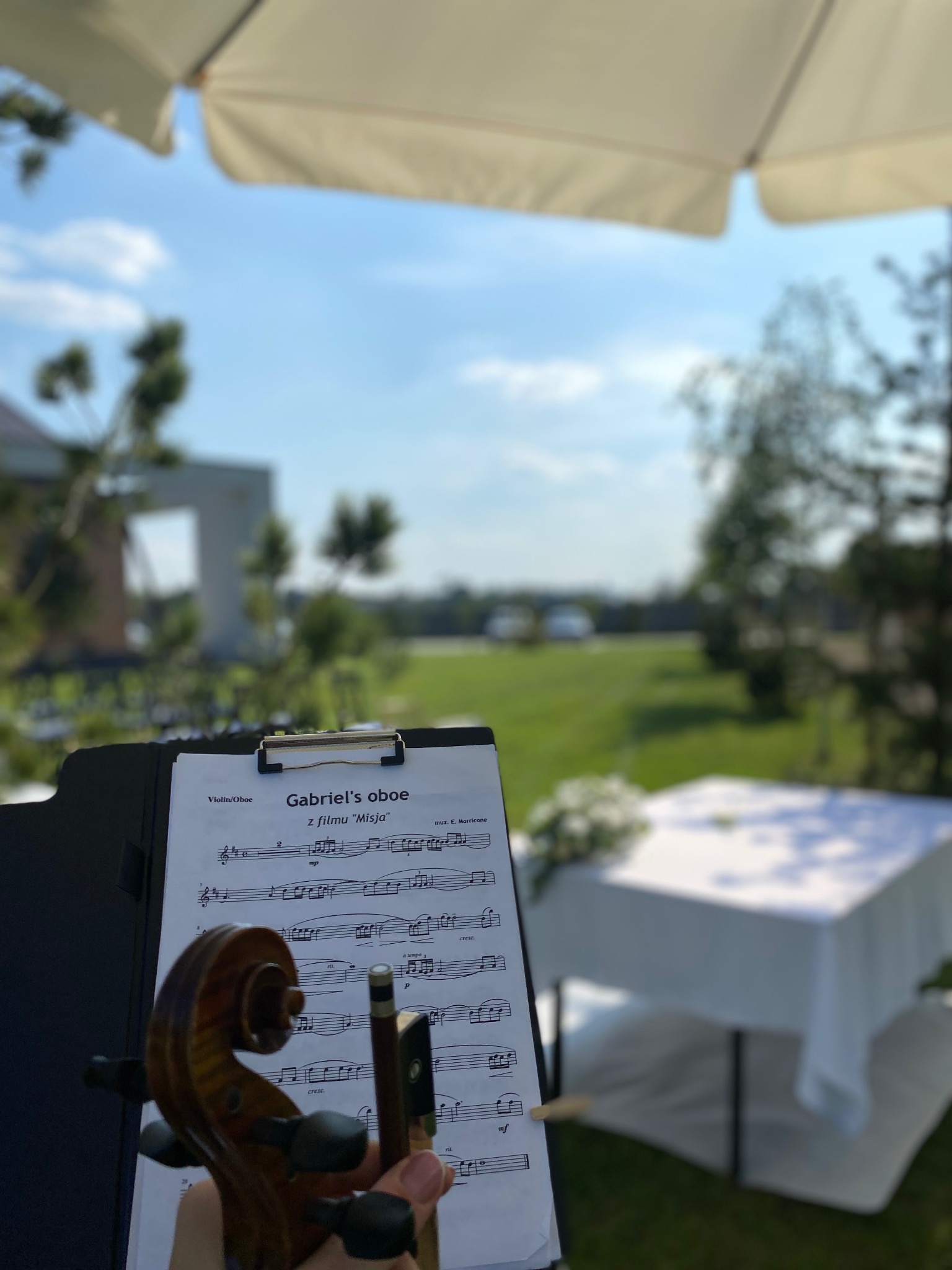 Skrzypce i nuty 'Gabriel's oboe' na tle plenerowej uroczystości. Biały obrus, zieleń trawy, błękit nieba. Idealne na ślub lub event.