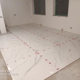 AM WORK SERVICES SPÓŁKA Z OGRANICZONĄ ODPOWIEDZIALNOŚCIĄ - Układanie białych płytek imitujących marmur na podłodze, z użyciem czerwonych krzyżyków dystansowych. Widoczne okna z zielonymi ramami i surowy stan wykończenia ścian.