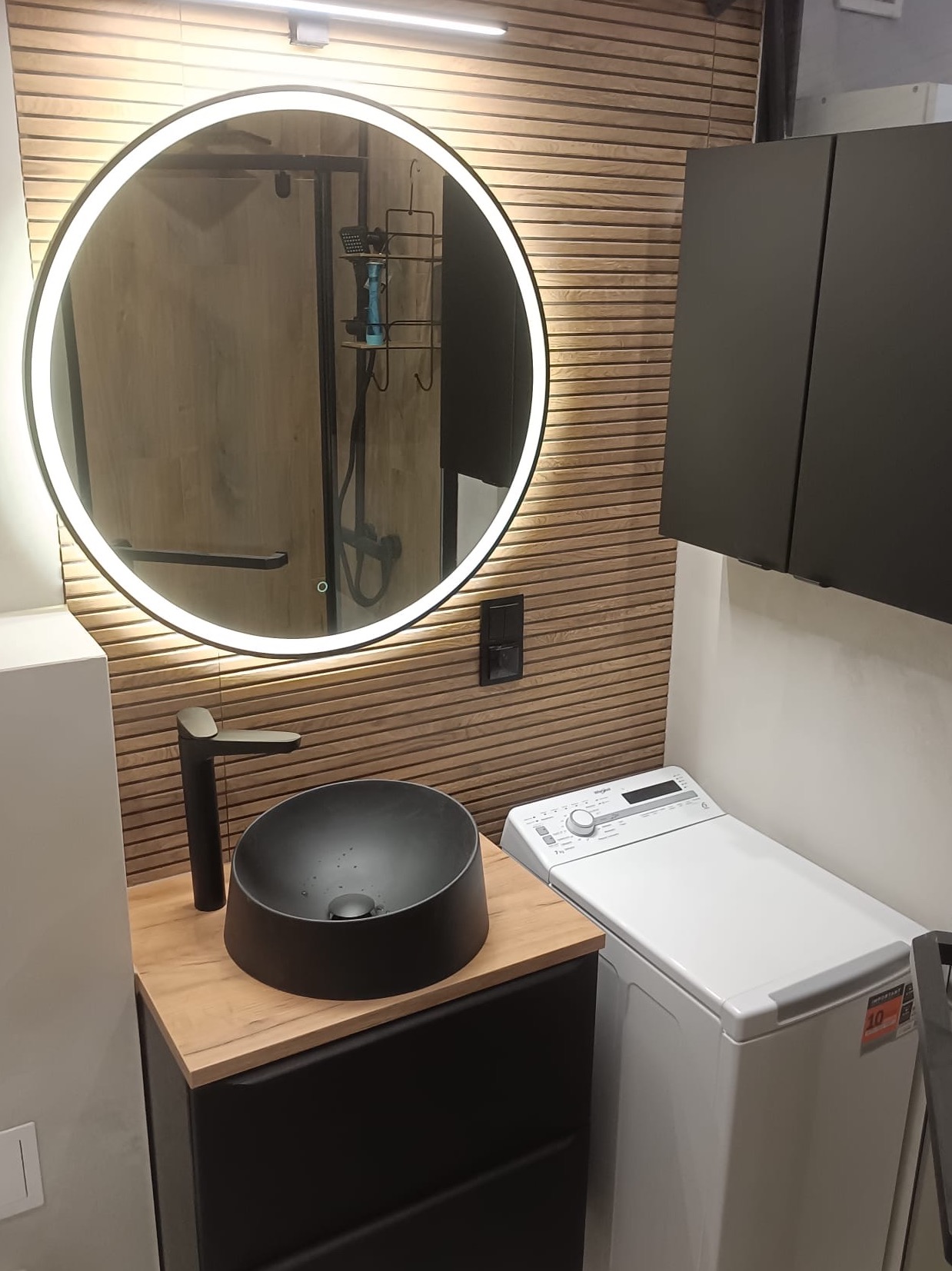 Nowoczesna łazienka z okrągłym lustrem LED, czarną umywalką nablatową i pralką. Ściana wykończona drewnopodobnymi panelami. Minimalistyczny design.