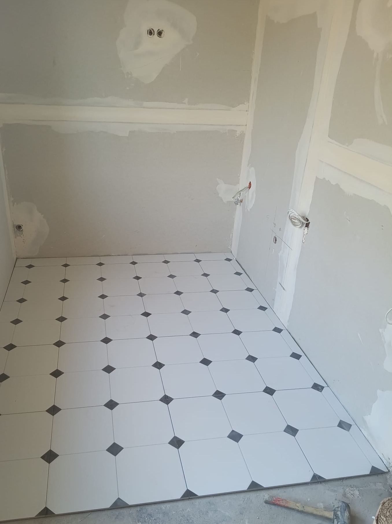 Biało-czarna podłoga z płytek w romby w trakcie remontu łazienki. Ściany w szpachli, widoczne kable. Młotek leży na podłodze. Ujęcie z góry.