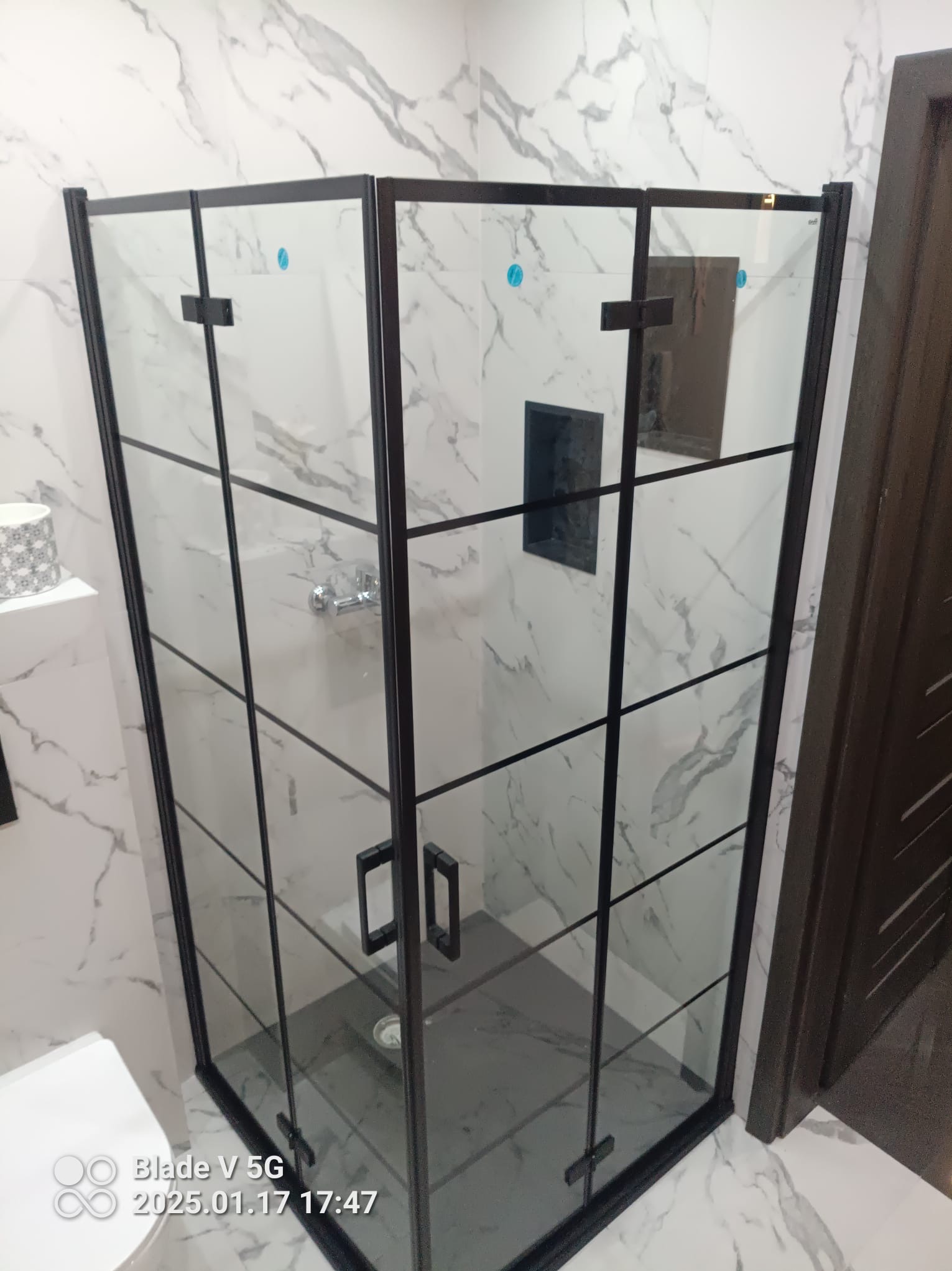 Nowoczesna kabina prysznicowa z czarnymi profilami na tle marmurowej ściany. Widoczny fragment toalety i drzwi. Minimalistyczny design, szklane panele.