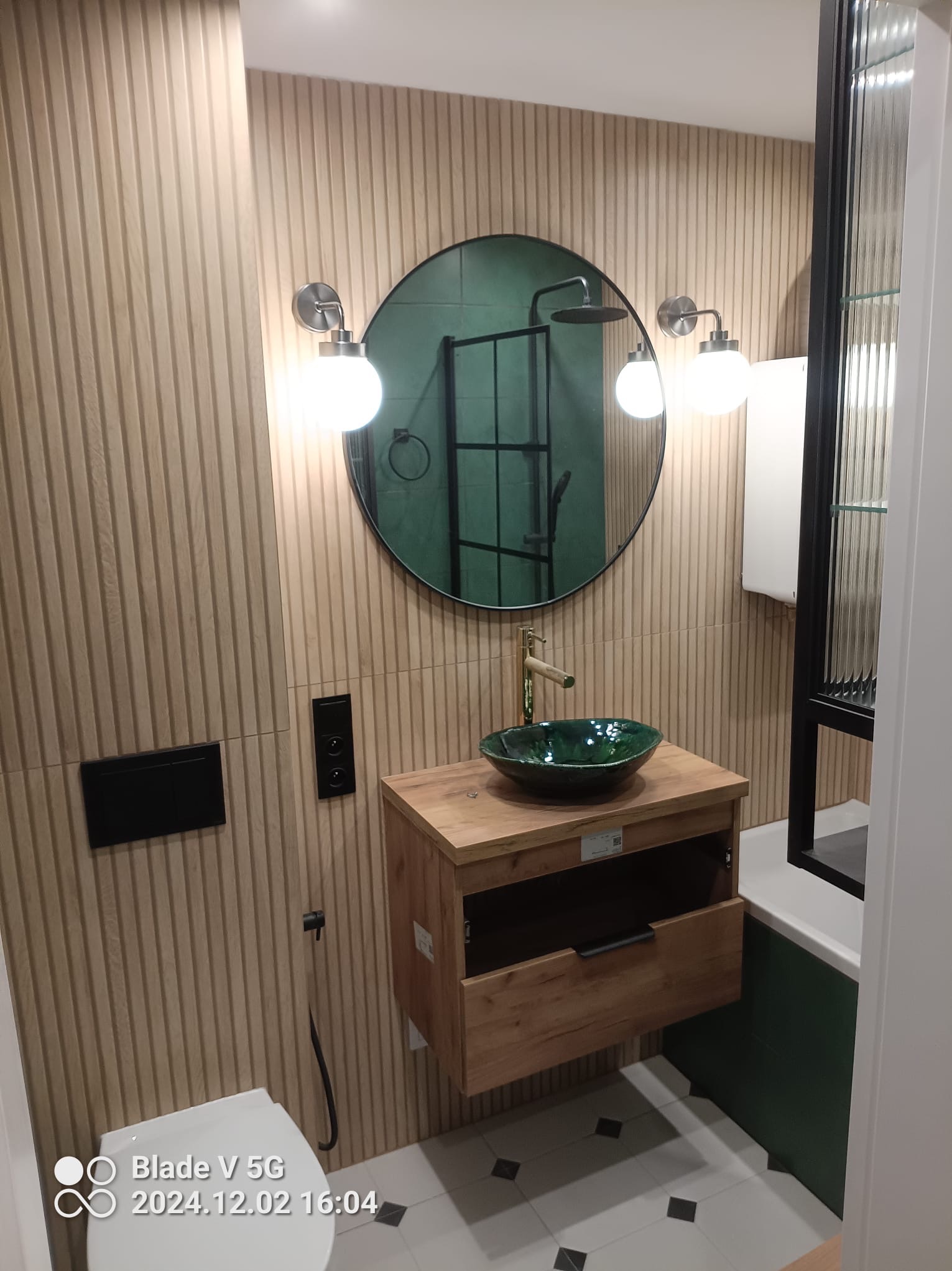 Nowoczesna łazienka z drewnianą ścianą, okrągłym lustrem i zieloną umywalką. Widoczna toaleta i szafka pod umywalką z otwartą szufladą. Minimalistyczny design.