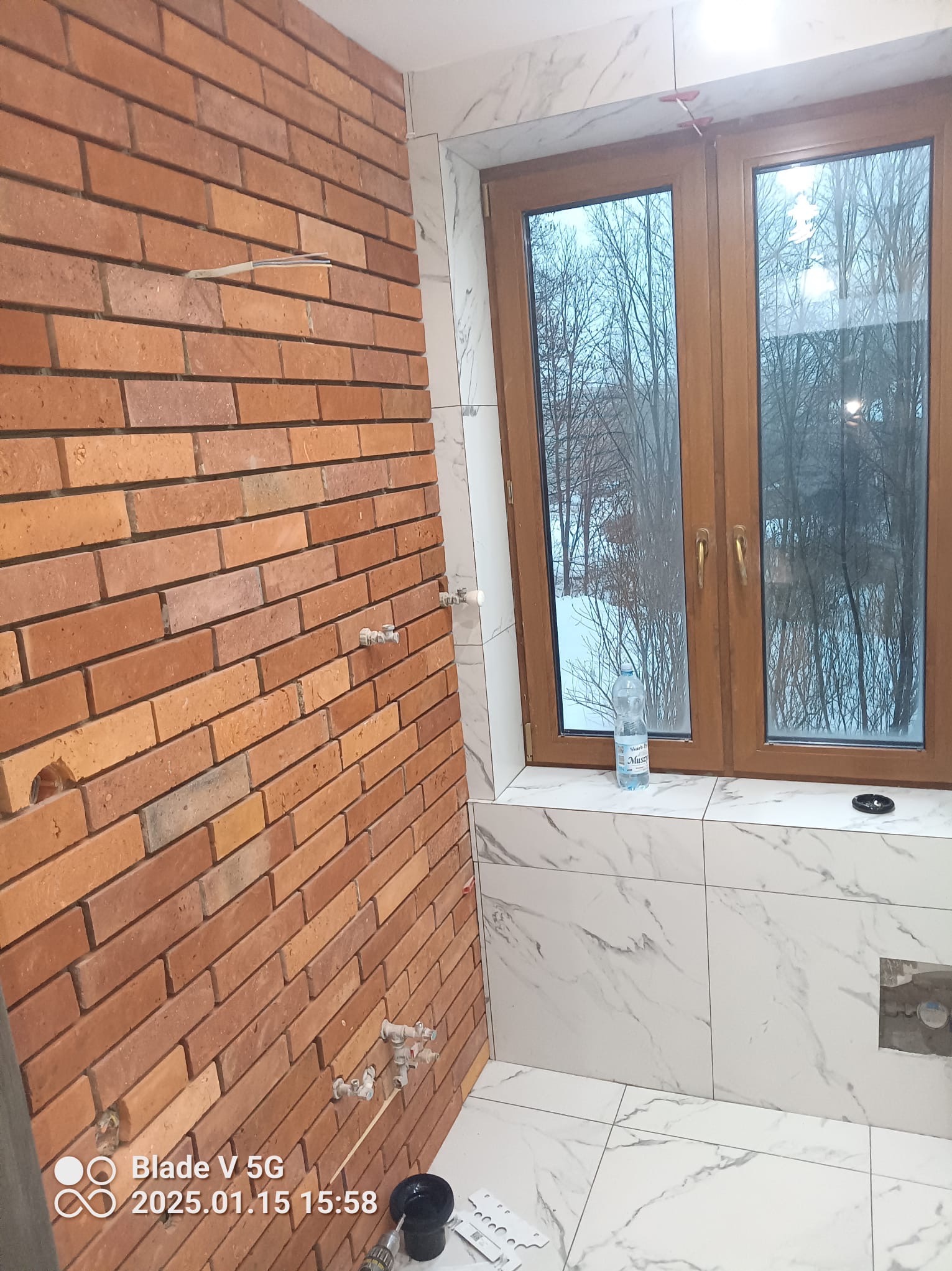 Łazienka w trakcie wykończenia: ściana z cegły klinkierowej z instalacją, białe płytki imitujące marmur, okno z widokiem na zimowy krajobraz. Widoczne narzędzia glazurnicze.