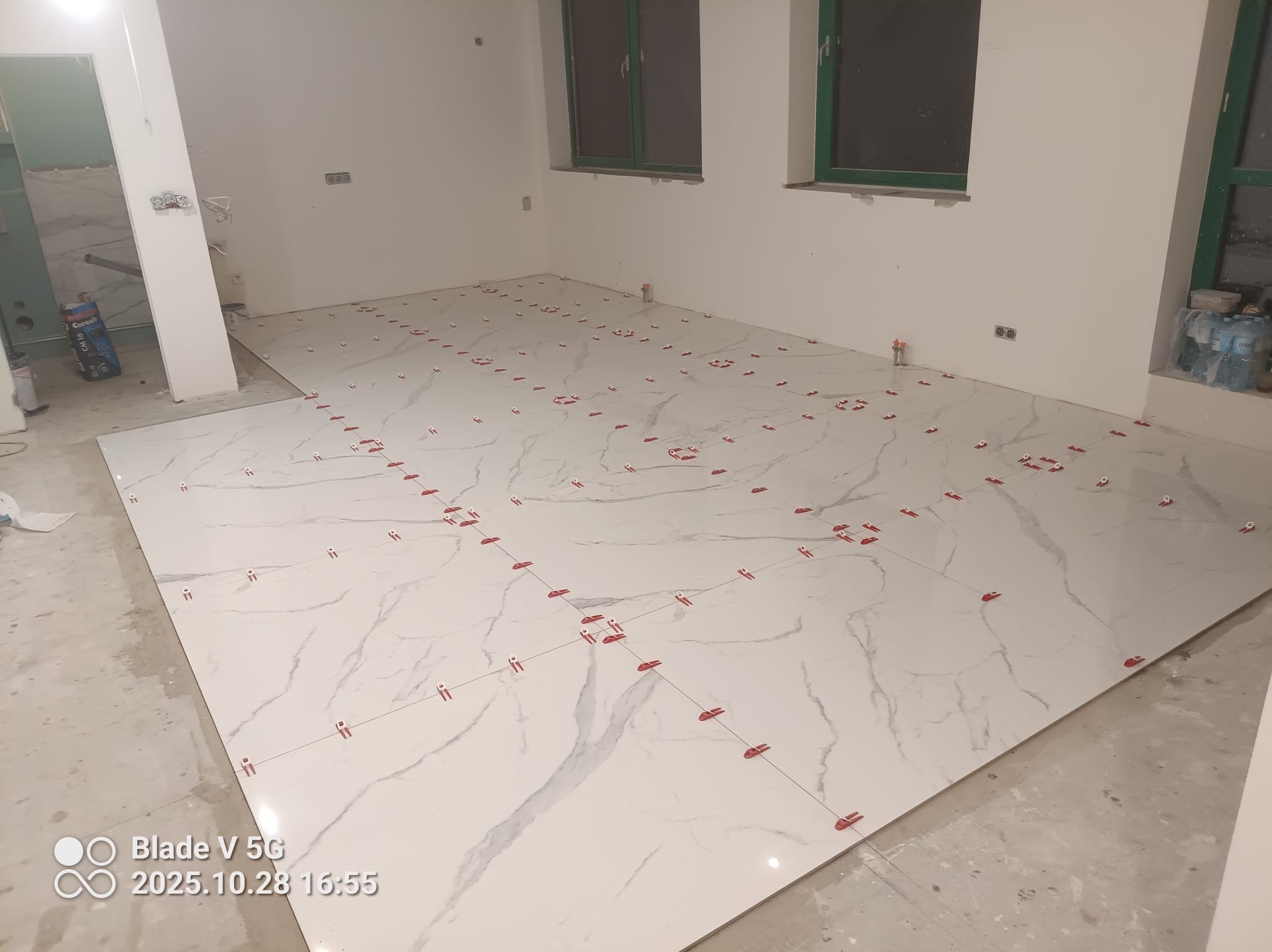 Układanie białych płytek imitujących marmur na podłodze, z użyciem czerwonych krzyżyków dystansowych. Widoczne okna z zielonymi ramami i surowy stan wykończenia ścian.