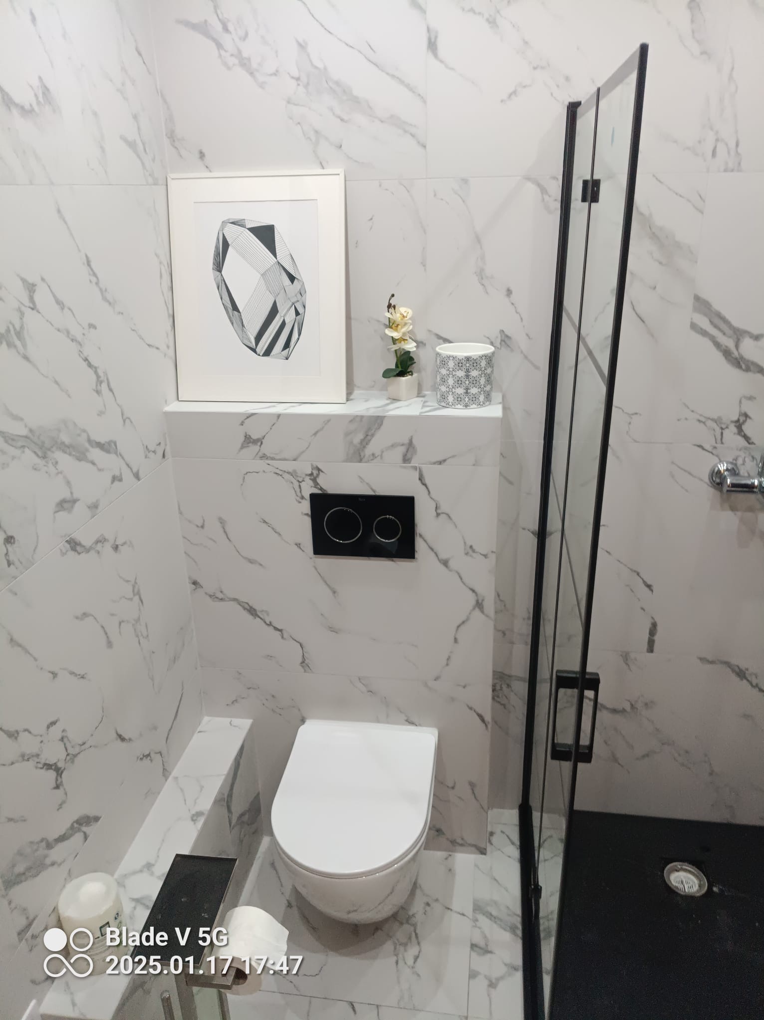 Biała łazienka z marmurowymi płytkami, wiszącą toaletą i czarną kabiną prysznicową. Na półce dekoracja i grafika w ramce. Nowoczesny design.