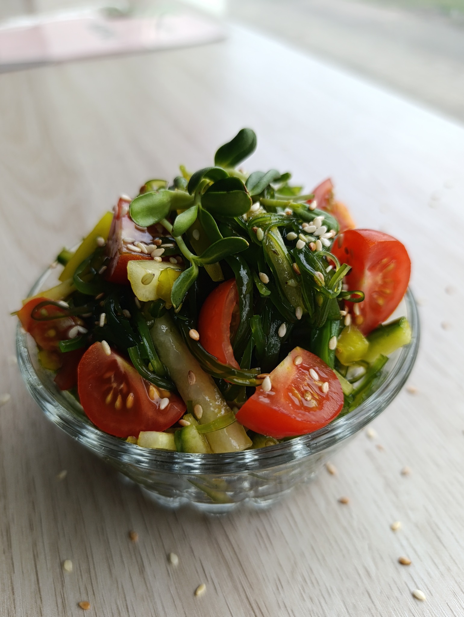 Szklana miseczka z sałatką z wodorostów, pomidorów i ogórków, posypana sezamem i kiełkami słonecznika, na jasnym drewnianym stole. Ujęcie z bliska.