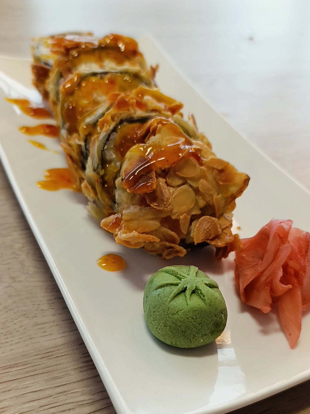 Sushi rolka w tempurze z migdałami, polana sosem, podana z wasabi i imbirem na białym talerzu. Nietypowa kompozycja smakowa, zbliżenie na detale.