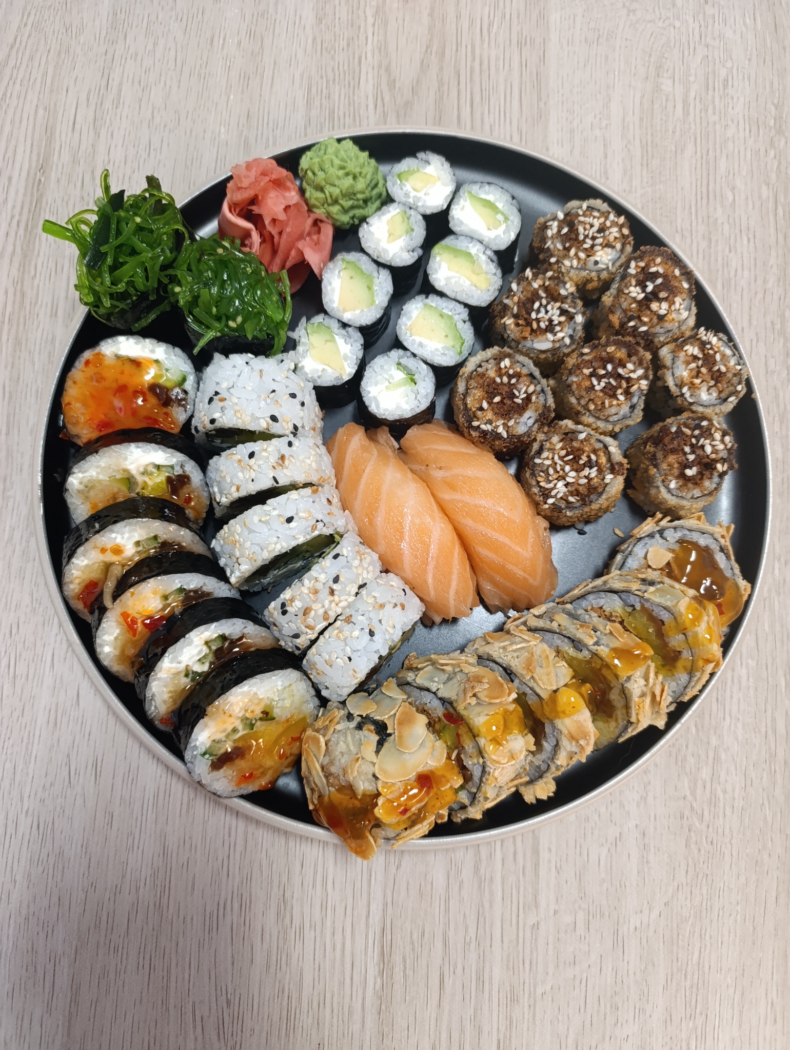 Zestaw sushi na czarnym talerzu: różnorodne rolki z łososiem, awokado, sezamem, tempura, algi wakame, imbir i wasabi. Elegancka kompozycja kulinarna.