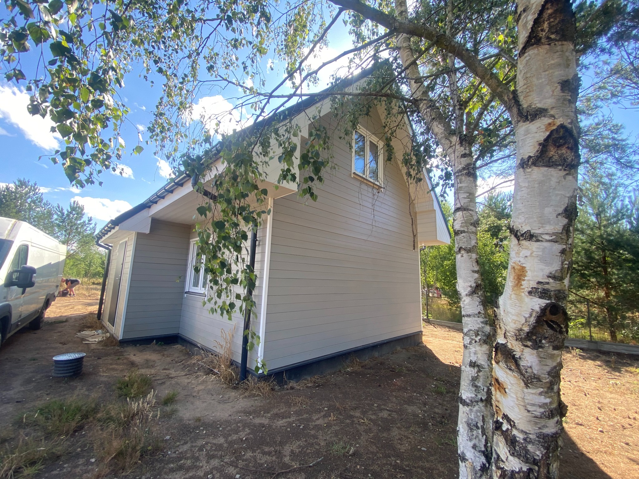 Gotowy, parterowy dom szkieletowy z szarą elewacją, widoczny z boku, z fragmentem białej brzozy na pierwszym planie i błękitnym niebem w tle. Realizacja Stare Wierzchowo.