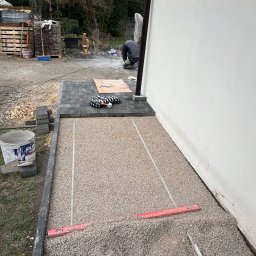 Renowo Dawid Sala - Układanie kostki brukowej przy ścianie budynku. Częściowo ułożona kostka, podsypka z piasku, poziomica i narzędzia. Prace brukarskie w toku, widoczne materiały budowlane.