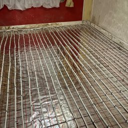 Renowo Dawid Sala - Ułożone rury ogrzewania podłogowego na folii izolacyjnej z siatką, widoczne fragmenty ścian w trakcie remontu, okno z firanką w tle.