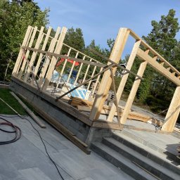 Renowo Dawid Sala - Drewniana konstrukcja szkieletowa budynku w trakcie budowy, widoczna na tle zieleni i błękitnego nieba. Elementy konstrukcyjne z jasnego drewna, fundament z szarej kostki.