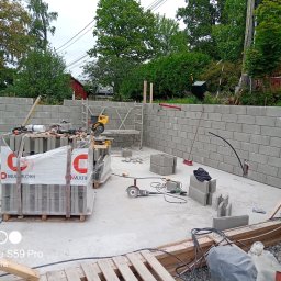 Renowo Dawid Sala - Wznoszenie ściany z szarych bloczków betonowych Multiblokk. Widoczne narzędzia murarskie, paleta z bloczkami i fragment krajobrazu z zielenią w tle.