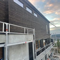 Renowo Dawid Sala - Elewacja domu z ciemnego drewna, częściowo otynkowana, z rusztowaniem. Widoczny pracownik na rusztowaniu i krajobraz w tle. Ujęcie z dołu, w pochmurny dzień.