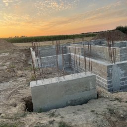 Renowo Dawid Sala - Wykopane fundamenty z betonowymi bloczkami i wystającymi prętami zbrojeniowymi na tle pola i zachodzącego słońca. Etap budowy domu.